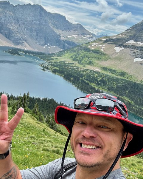Mike Saur, smiling on a grassy alpine ridge above Hidden Lake in Glacier National Park, with the blue lake and rocky peaks behind him